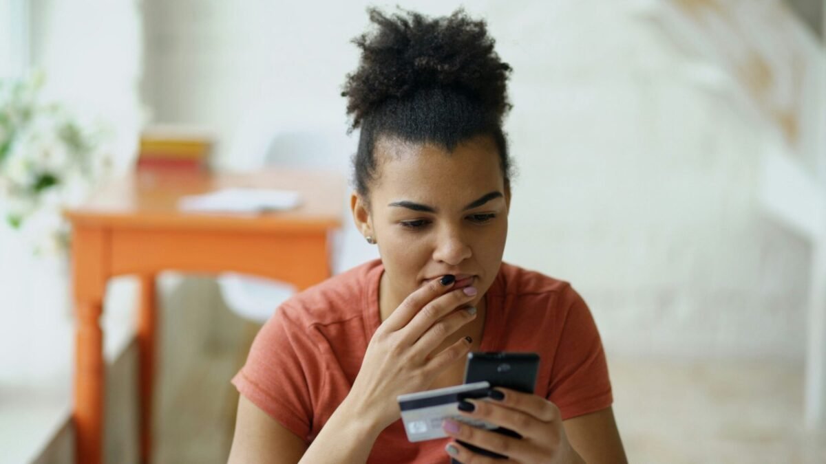 Mulher preocupada segurando cartão de crédito e celular, refletindo sobre suas dívidas e gastos.