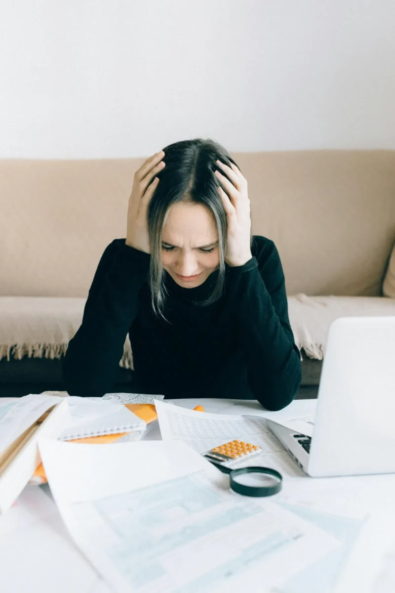 Mulher com as mãos na cabeça, visivelmente estressada diante de uma mesa cheia de documentos, calculadora e notebook aberto.