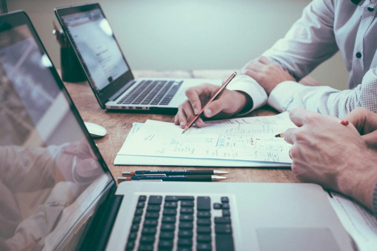 Pessoas analisando documentos financeiros e fazendo cálculos em uma mesa com notebooks e calculadora