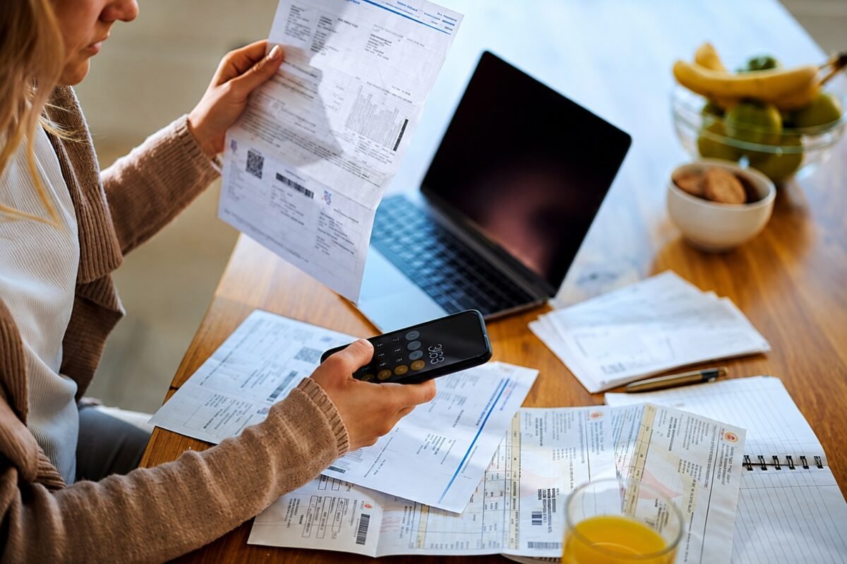 Mulher analisando contas e documentos financeiros enquanto usa uma calculadora no celular, com notebook sobre a mesa.